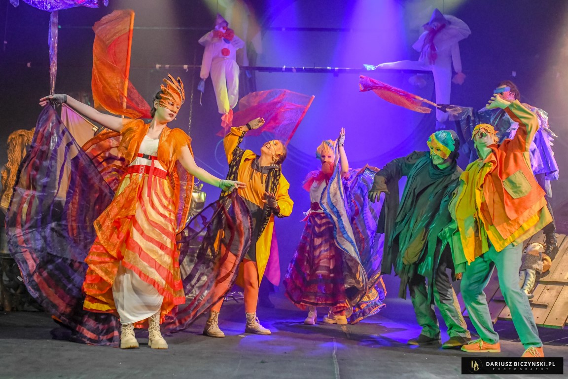 Grupa aktorów w strojach w kolorach tęczy tańczy w grupie. Wykonują dynamiczne ruchy machając atrybutami z materiałów nad głowami.